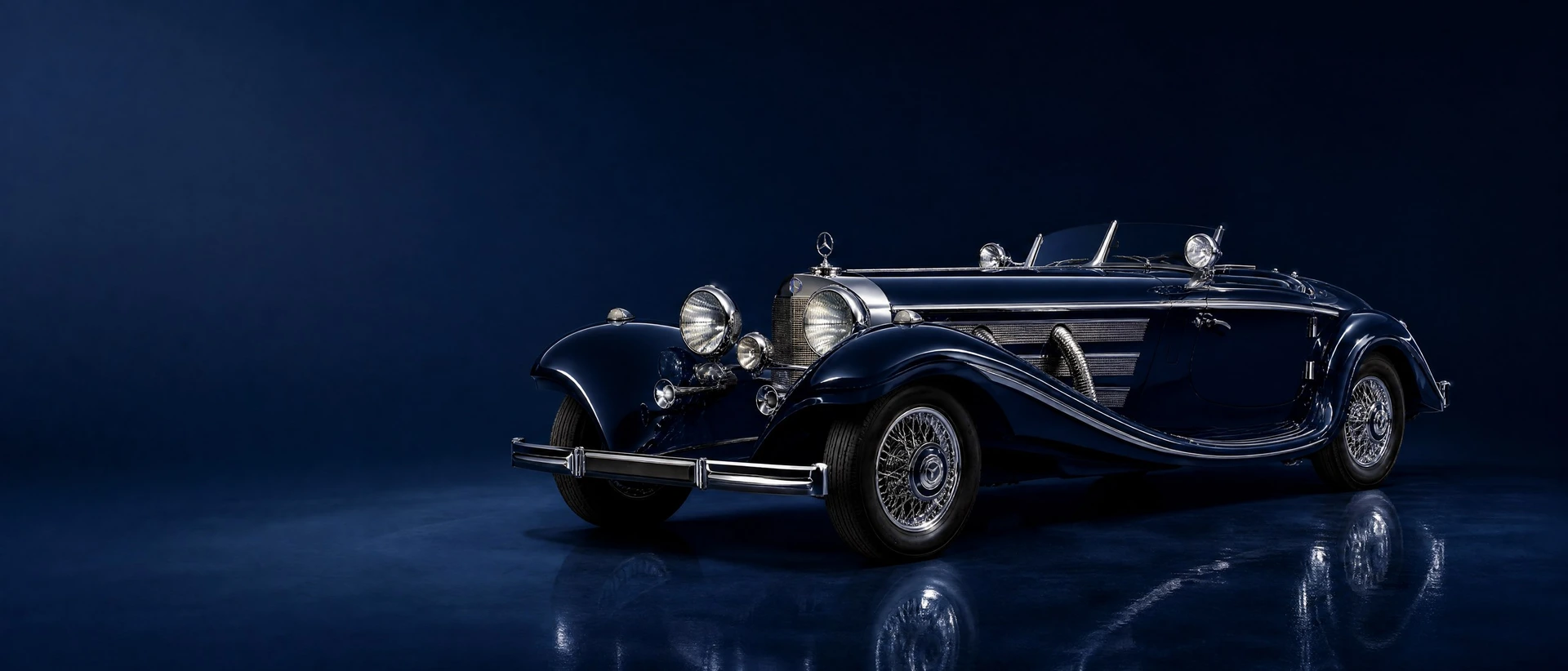 A midnight-blue Mercedes-Benz 500 K Spezial-Roadster with chrome exhaust pipes, front three-quarter view on a deep midnight-blue field in soft evening light