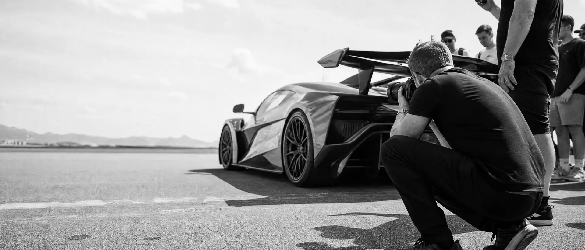 A photographer crouched low on the tarmac shooting a modern hypercar, spotters watching, at a daylight tour stop