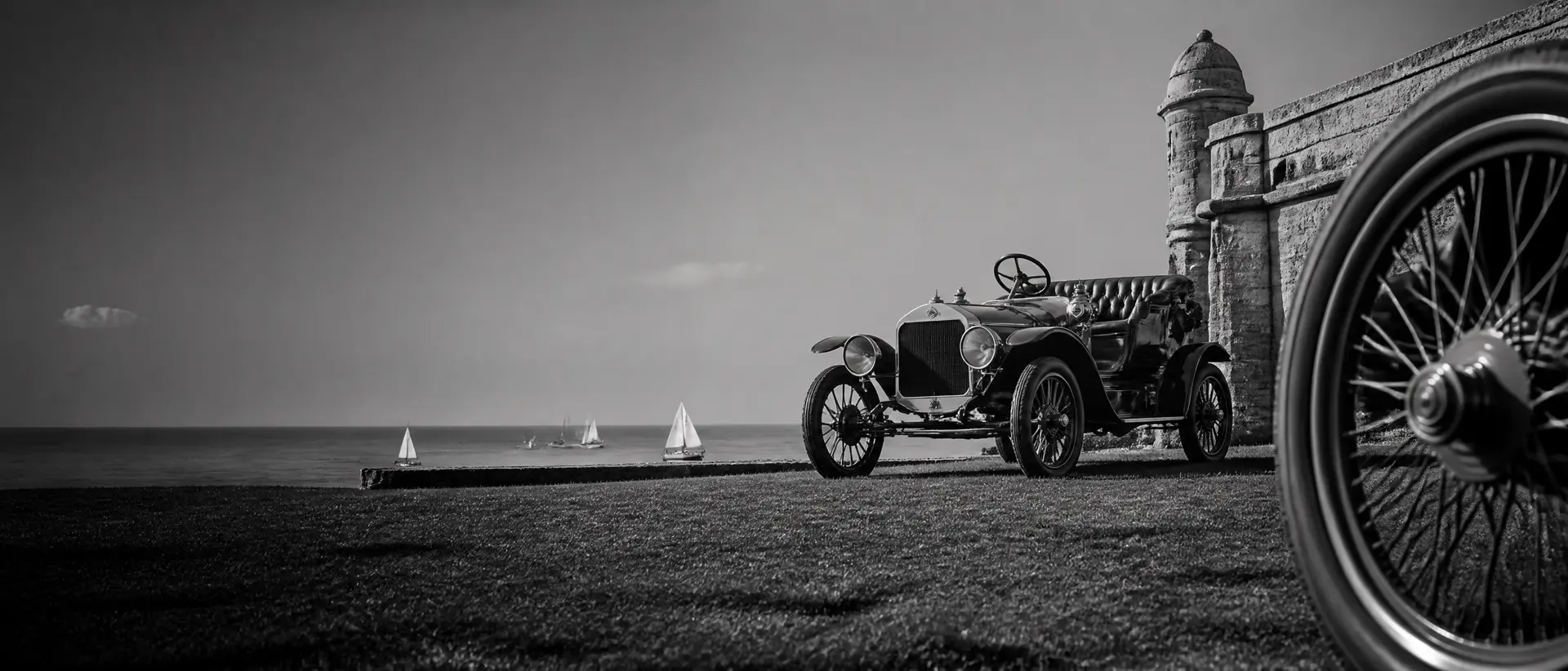 Black-and-white image of a brass-era automobile on a fortress green, sailboats monochrome on the bay behind, autumn light, a spoked wheel cropped hard at the frame’s edge