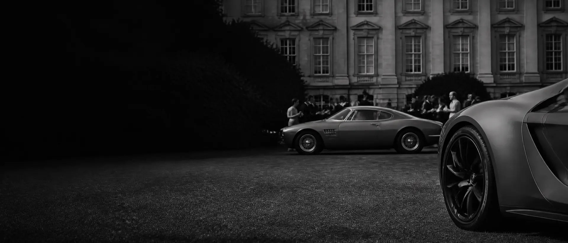 Black-and-white composition of a coachbuilt classic on a palace lawn, baroque stone façade soft behind, champagne guests out of focus, a modern hypercar’s wheel cropped at the right edge