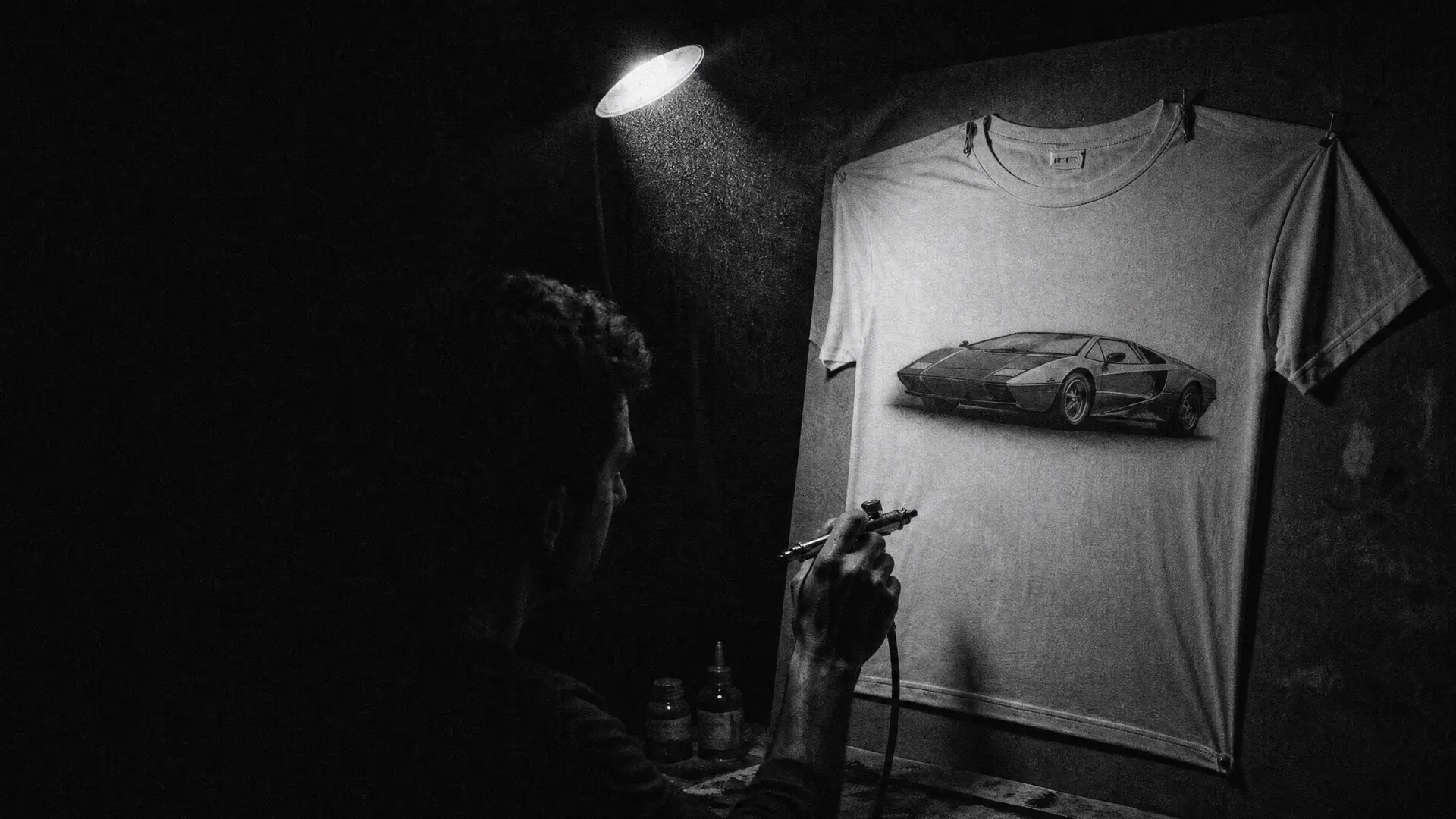 Art plate: a hand airbrushing a wedge supercar onto a pinned T-shirt under one bare lamp