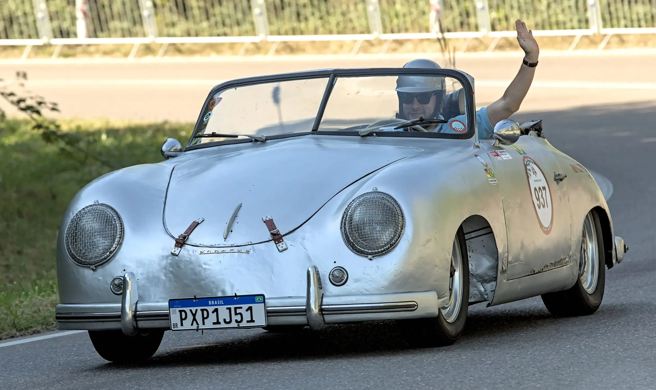 A silver Porsche 356 pre-A at speed, its driver waving