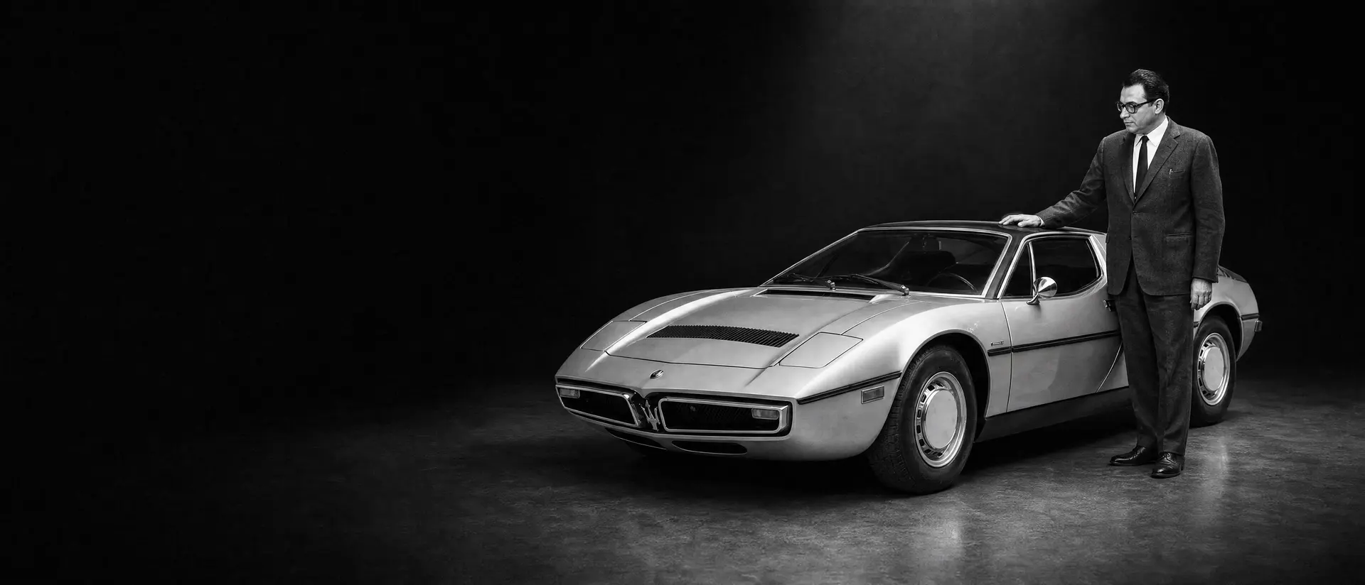 Giulio Alfieri in a dark period jacket standing beside a Maserati Bora under a single spotlight
