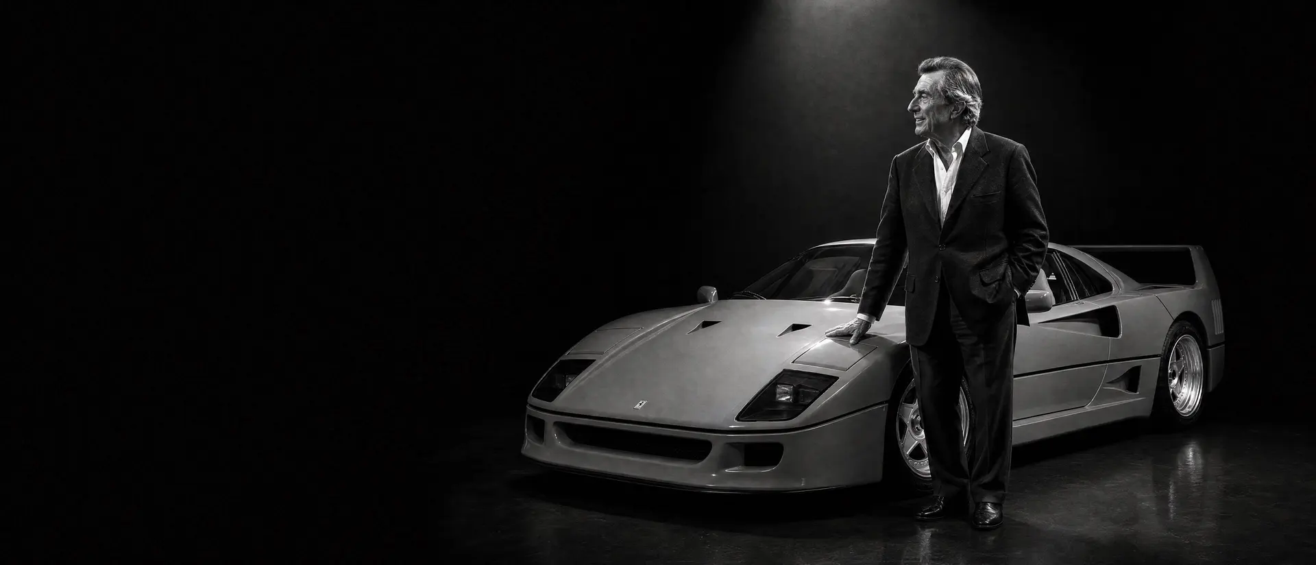 Leonardo Fioravanti in a tailored dark jacket standing beside a Ferrari F40 under a single overhead spotlight, his hand resting on the front wing